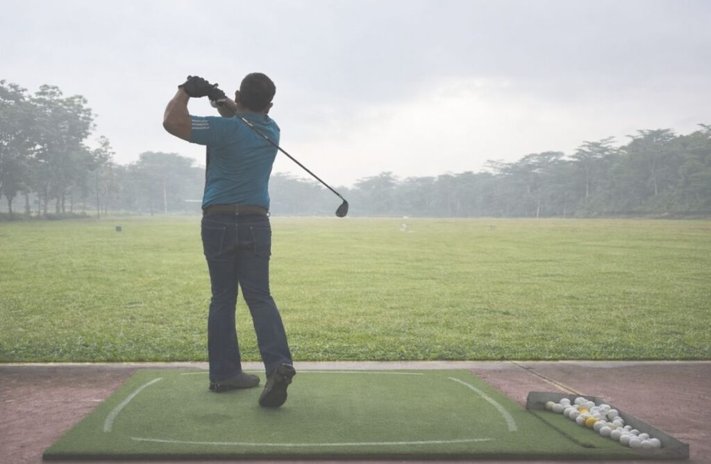 Pria mengayun stik golf di driving range berkabut dengan posisi follow through penuh fokus.