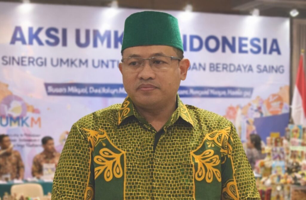 Seorang pria mengenakan peci hijau dan kemeja batik hijau bermotif kuning berdiri dengan latar belakang banner acara bertema strategi dan pengembangan, tampak formal dan serius.
