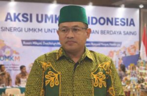 Seorang pria mengenakan peci hijau dan kemeja batik hijau bermotif kuning berdiri dengan latar belakang banner acara bertema strategi dan pengembangan, tampak formal dan serius.